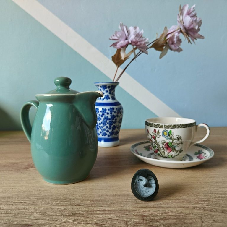 A green Denby coffee or tea pot in the foreground, with a blue and white small vase in the background and an Indian Tree tea cup and saucer