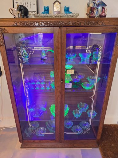 Display cabinet showcasing glowing uranium glassware and decorative items under UV light.